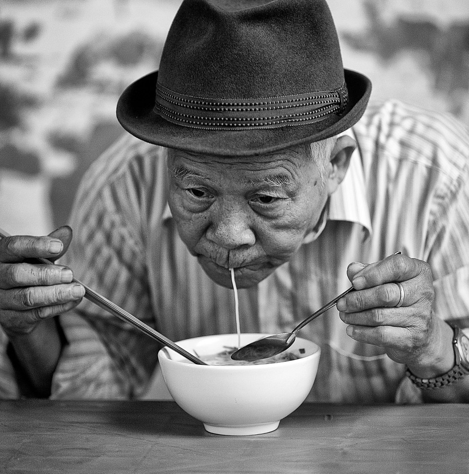En: An old man eating a noodle. Eo: Maljuna viro manĝanta nudelon.